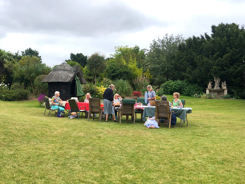 Ragged Life rag rug coffee morning showing a group of people sitting at tables in a garden setting with greenery and a thatched-roof structure.